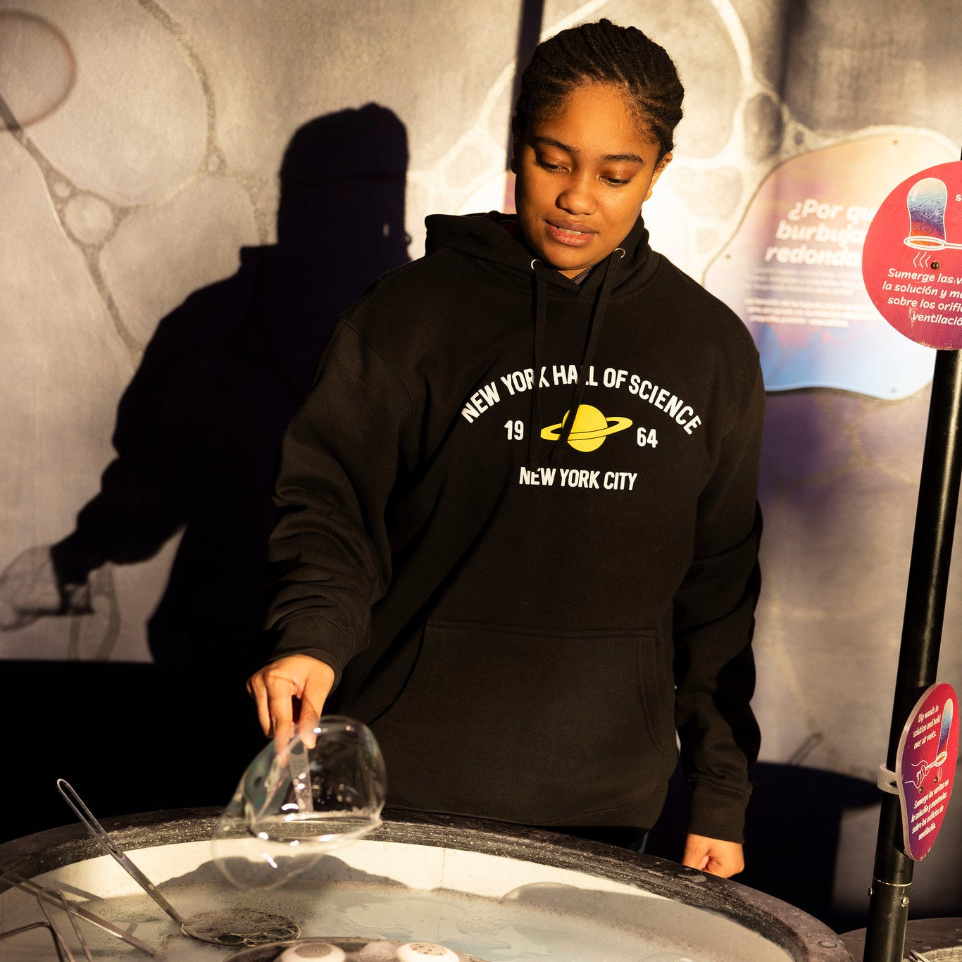 Woman wearing black NYSCI branded sweatshirt with yellow Saturn graphic at a display in the museum