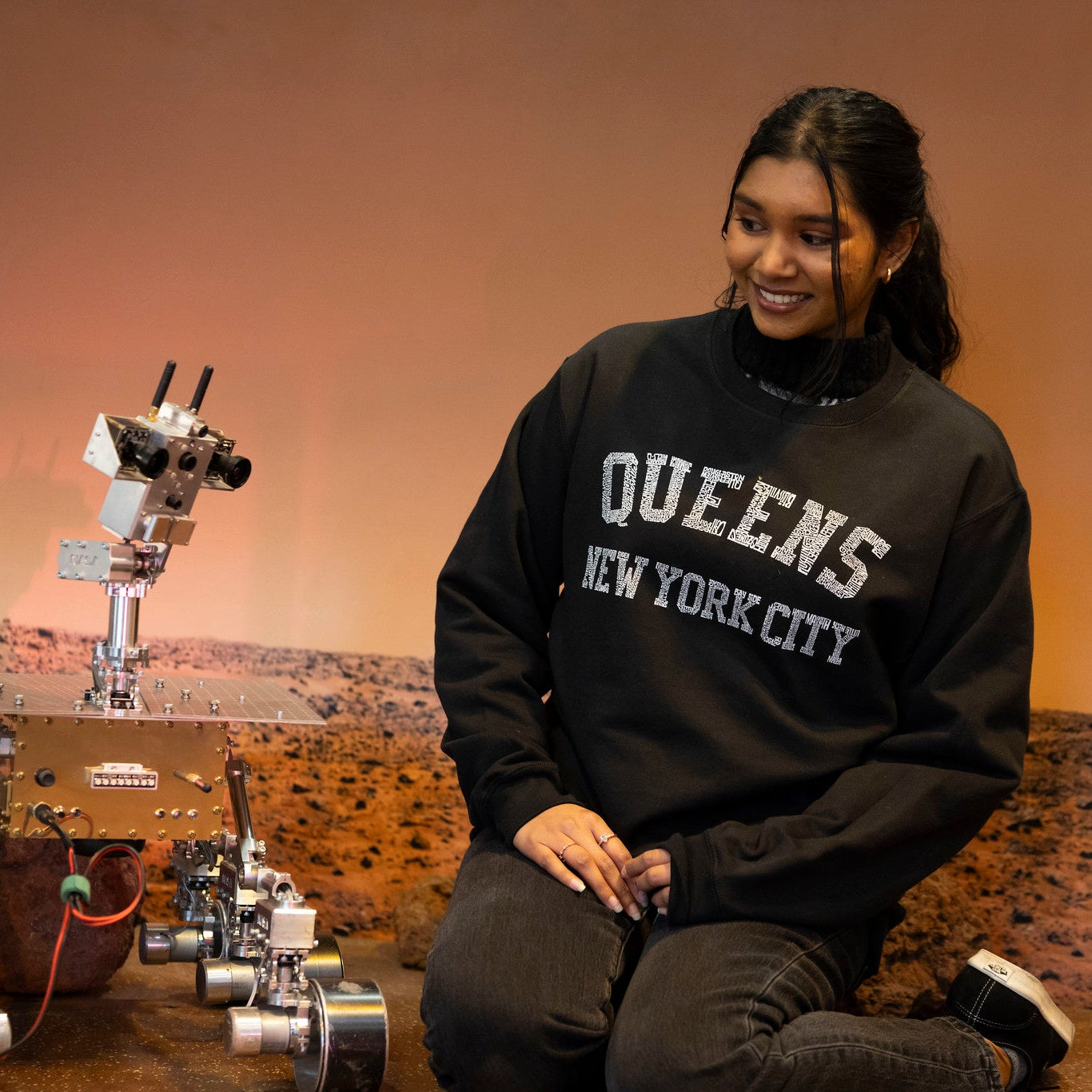 Woman wearing "Queens New York City" black sweatshirt sitting in a Mars Rover display at the museum