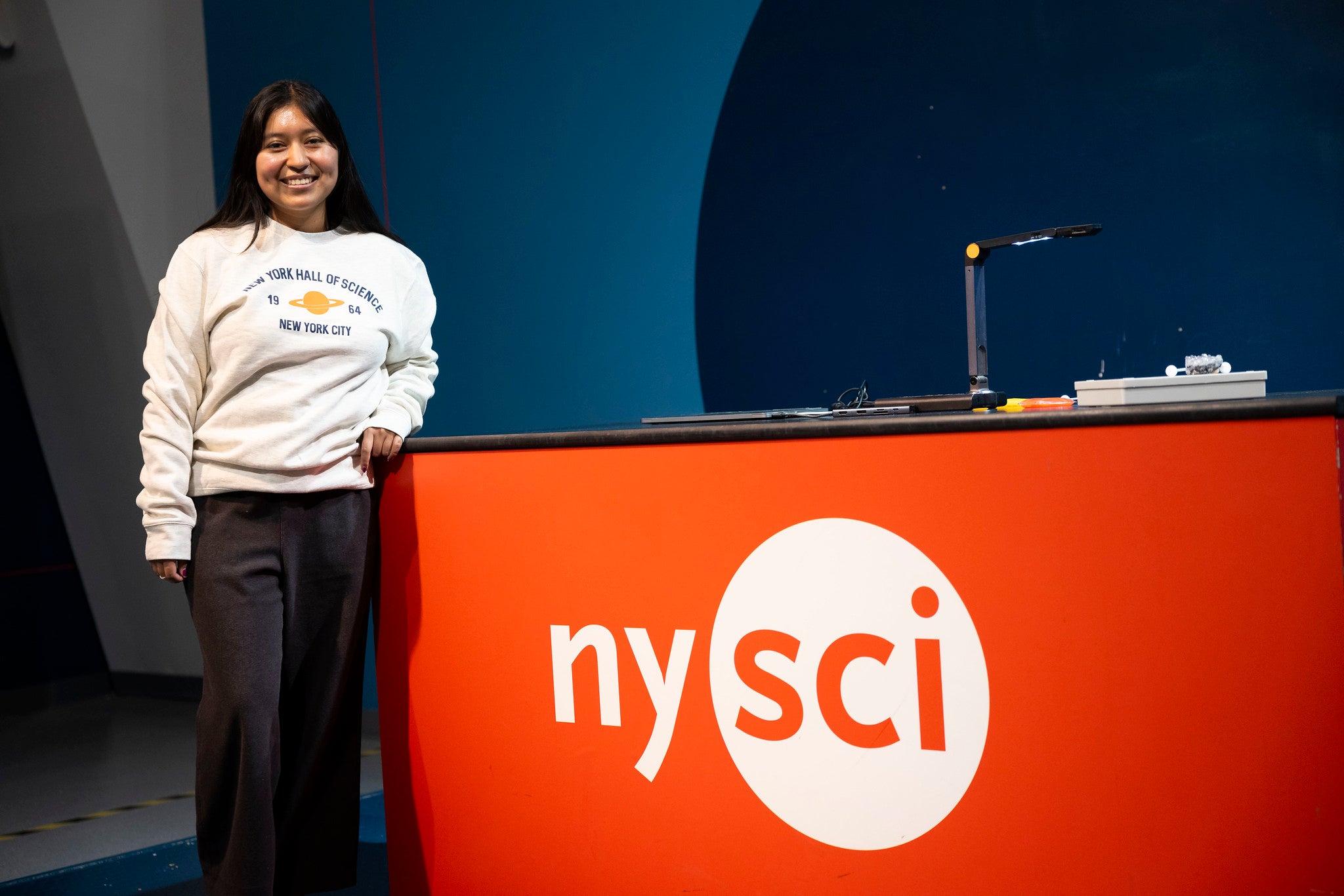 Beige sweatshirt on a girl at a NYSCI welcome desk with words NY Hall of Science, NYC 1964 and a graphic of Saturn