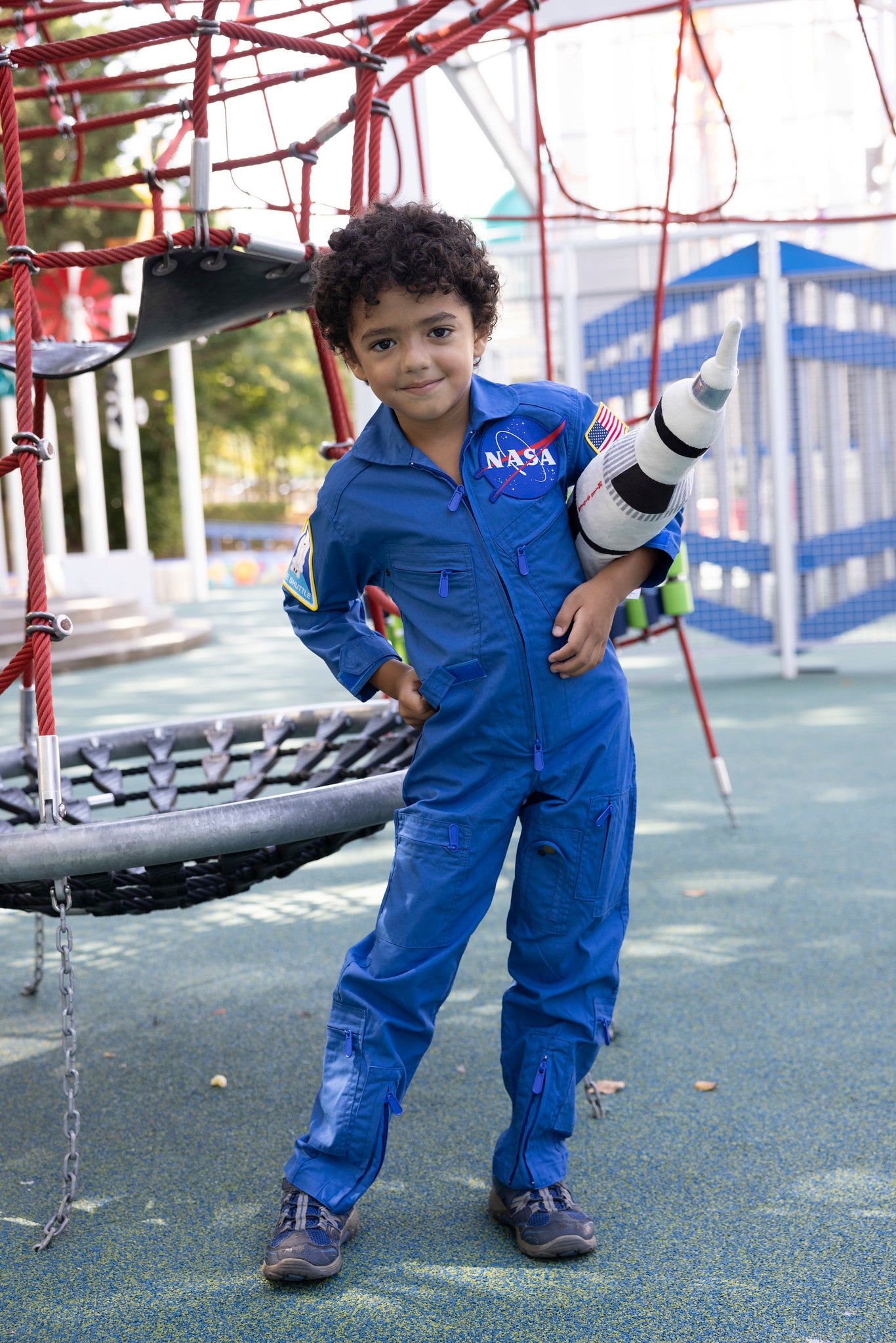 Child in front of a playground who is wear a blue coverall spacesuit, they are holding a space ship and have their hand on their hip. The coverall has the NASA meatball on the front.