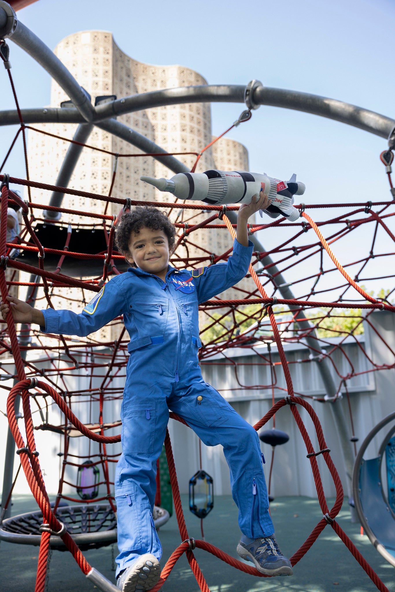 Child in front of a playground who is wear a blue coverall spacesuit, they are holding a space ship in the air as they hold onto the rope. The coverall has the NASA meatball on the front.