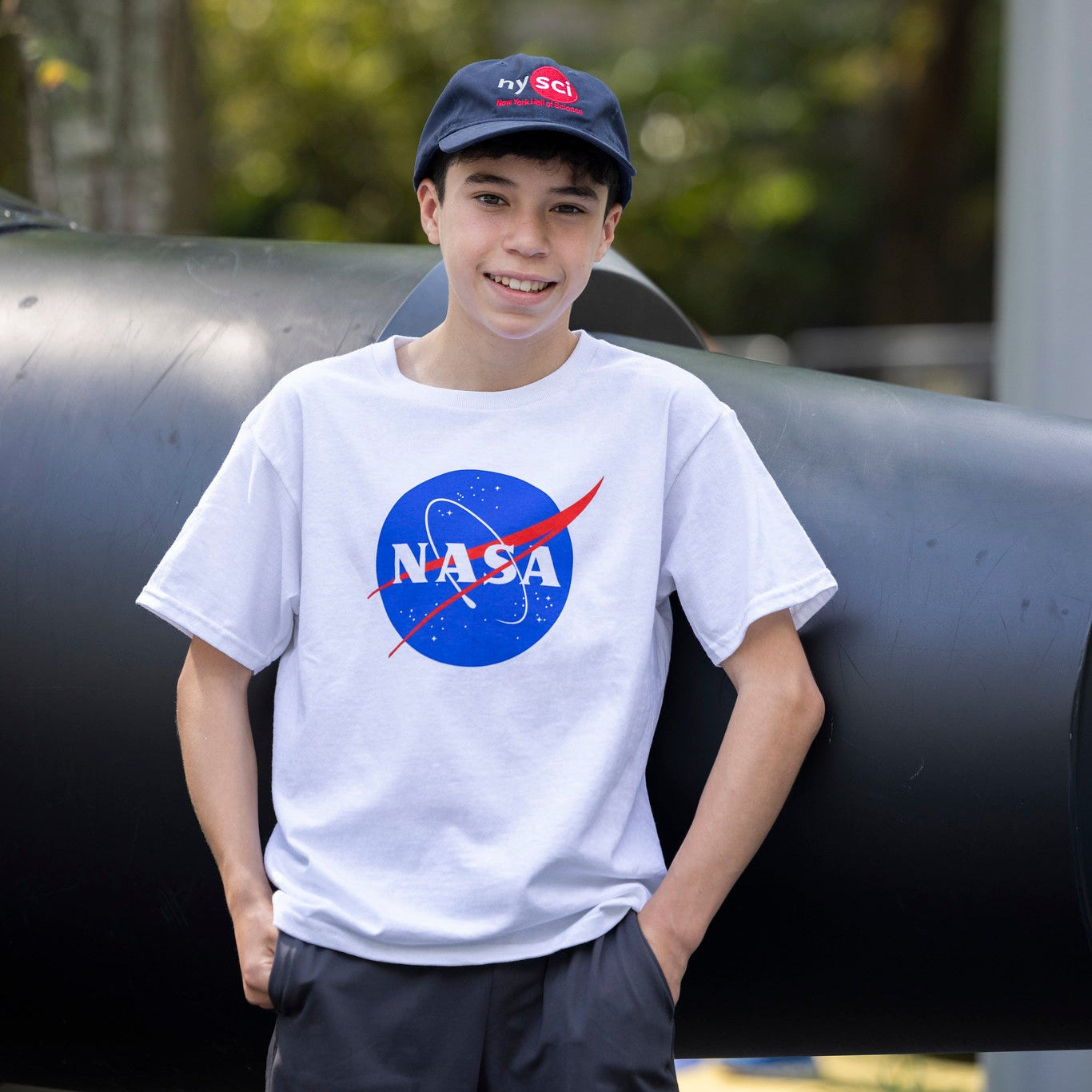 Child posing wearing white NASA t-shirt and nysci baseball cap outside