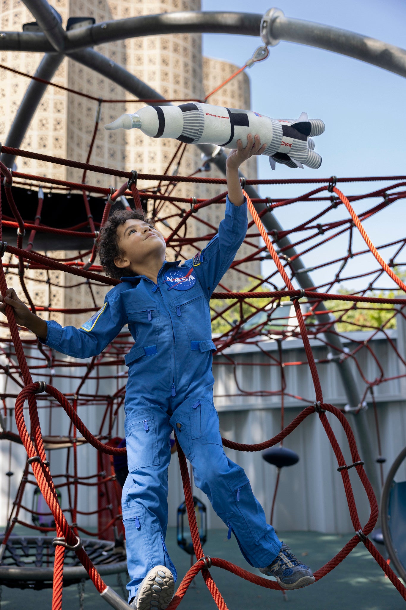 Child in front of a playground who is wear a blue coverall spacesuit, they are holding a space ship in the air as they hold onto the rope. The coverall has the NASA meatball on the front.