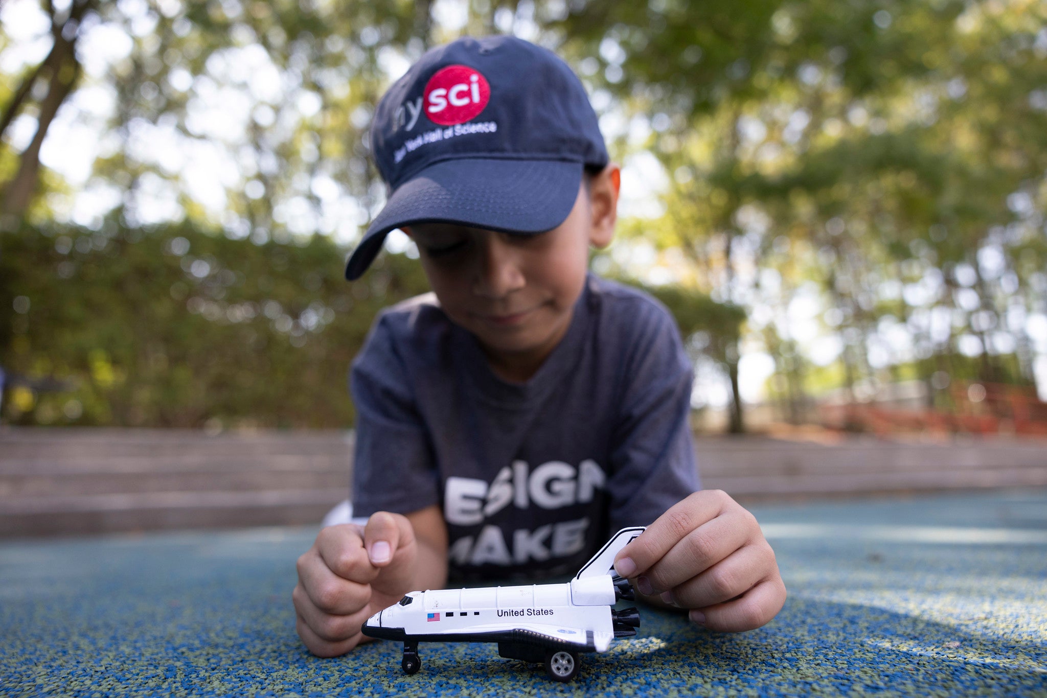 A child laying on his stomach on the ground with a baseball cap which says NYSCI. He is playing with a spaceship toy.
