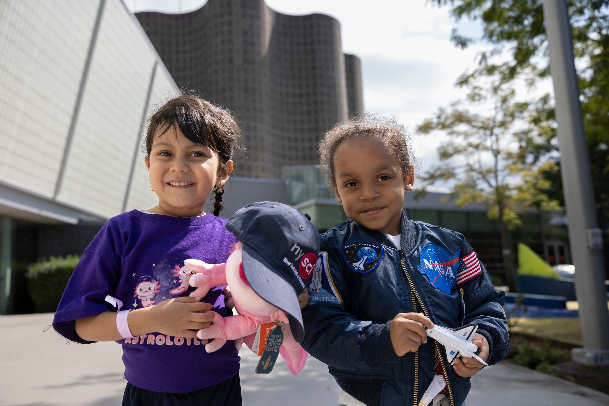 two young children wearing NASA and nysci branded t-shirts and jackets in front of museum buildings
