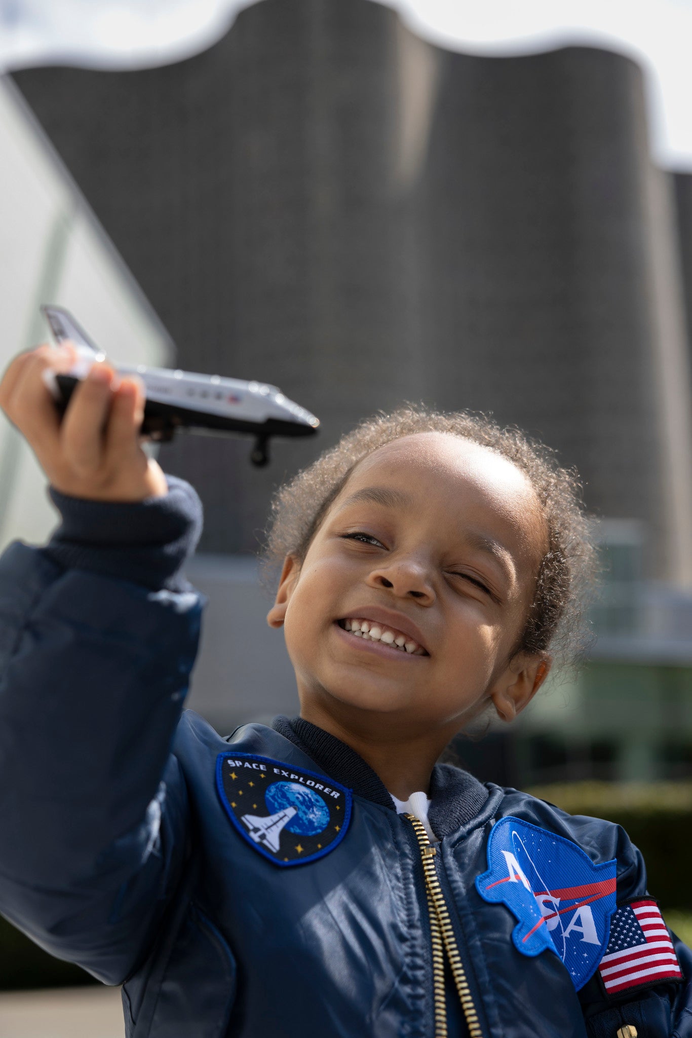young girl smiling and playing with a space shuttle toy wearing a NASA jacket