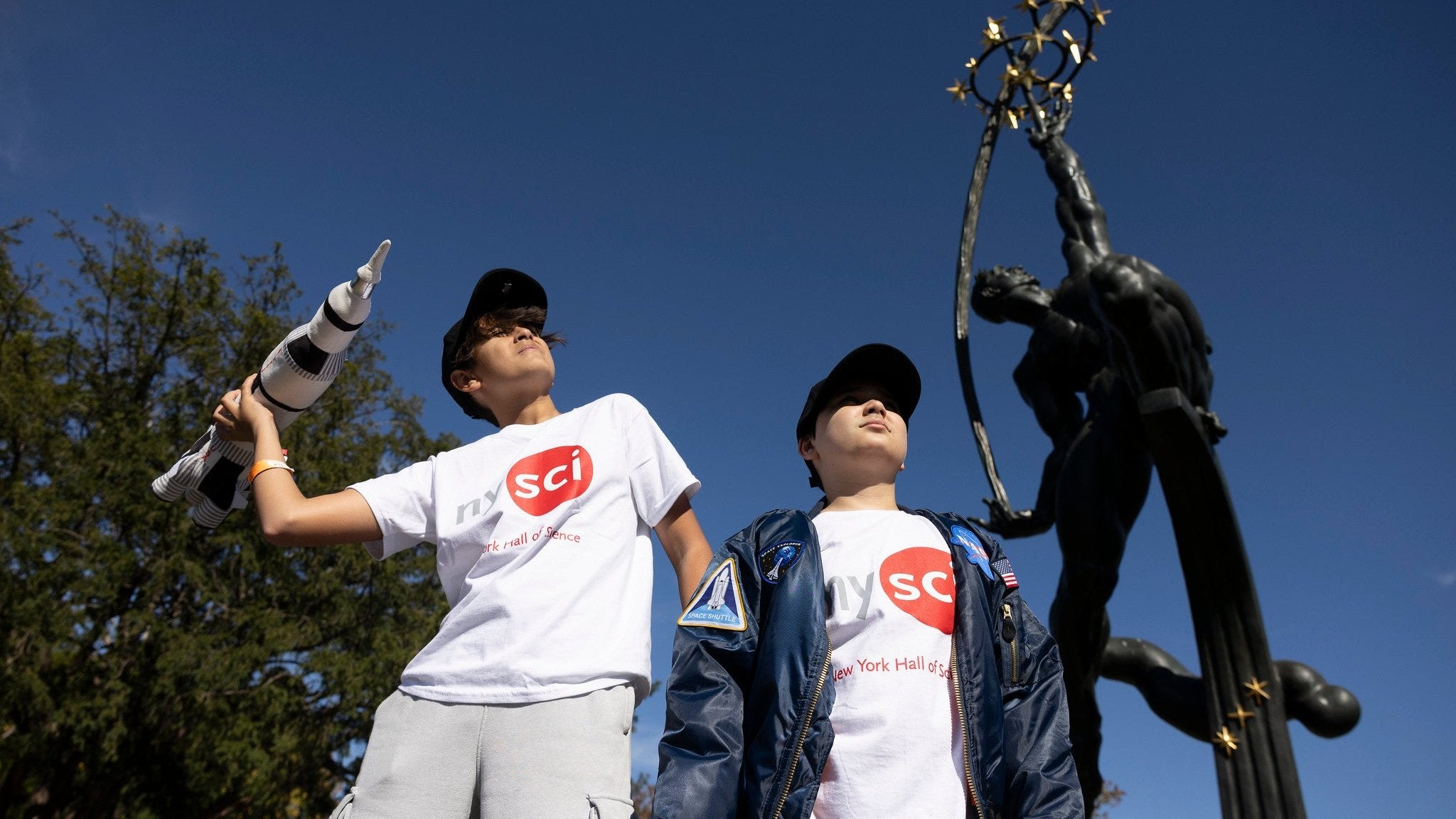 two children wearing white nysci t-shirts outside in the museum park holding a plush NASA rocket
