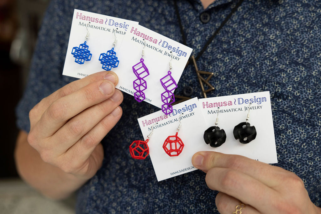 Hands holding Hanusa Design mathematical jewelry cards with various earrings.