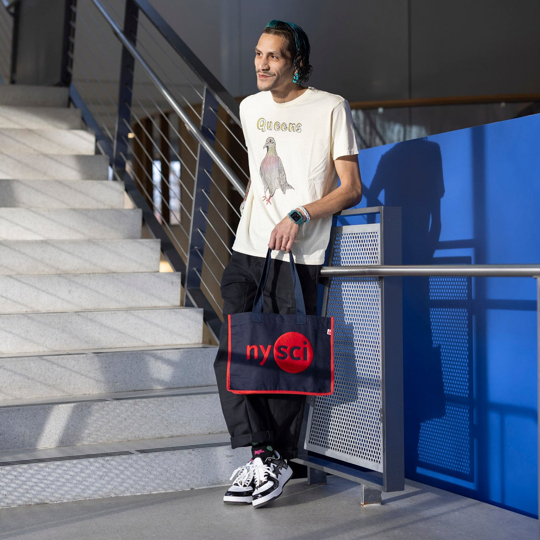 Man looking away from camera, wearing a queens pigeon Tshirt, and holding a Navy tote bag with Nysci logo