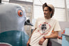 Man wearing off white tshirt with NYSCI text in red and astronaut graphic, standing inside