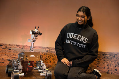 Woman wearing &quot;Queens New York City&quot; black sweatshirt sitting in a Mars Rover display at the museum