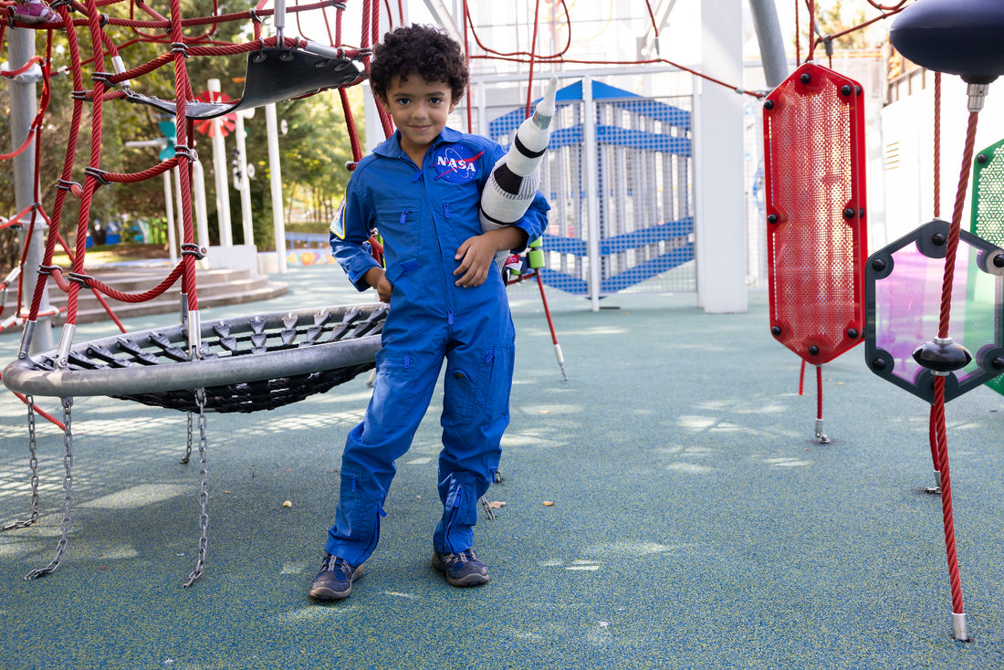 Child in front of a playground who is wear a blue coverall spacesuit, they are holding a space ship and have their hand on their hip. The coverall has the NASA meatball on the front.