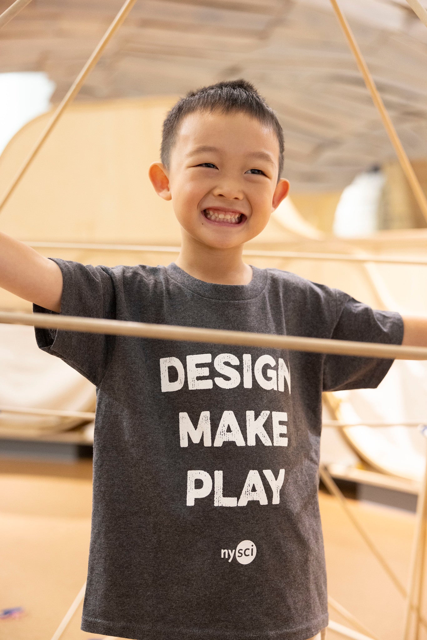 A child smiling at the camera with a grey t-shirt which says DESIGN MAKE PLAY.