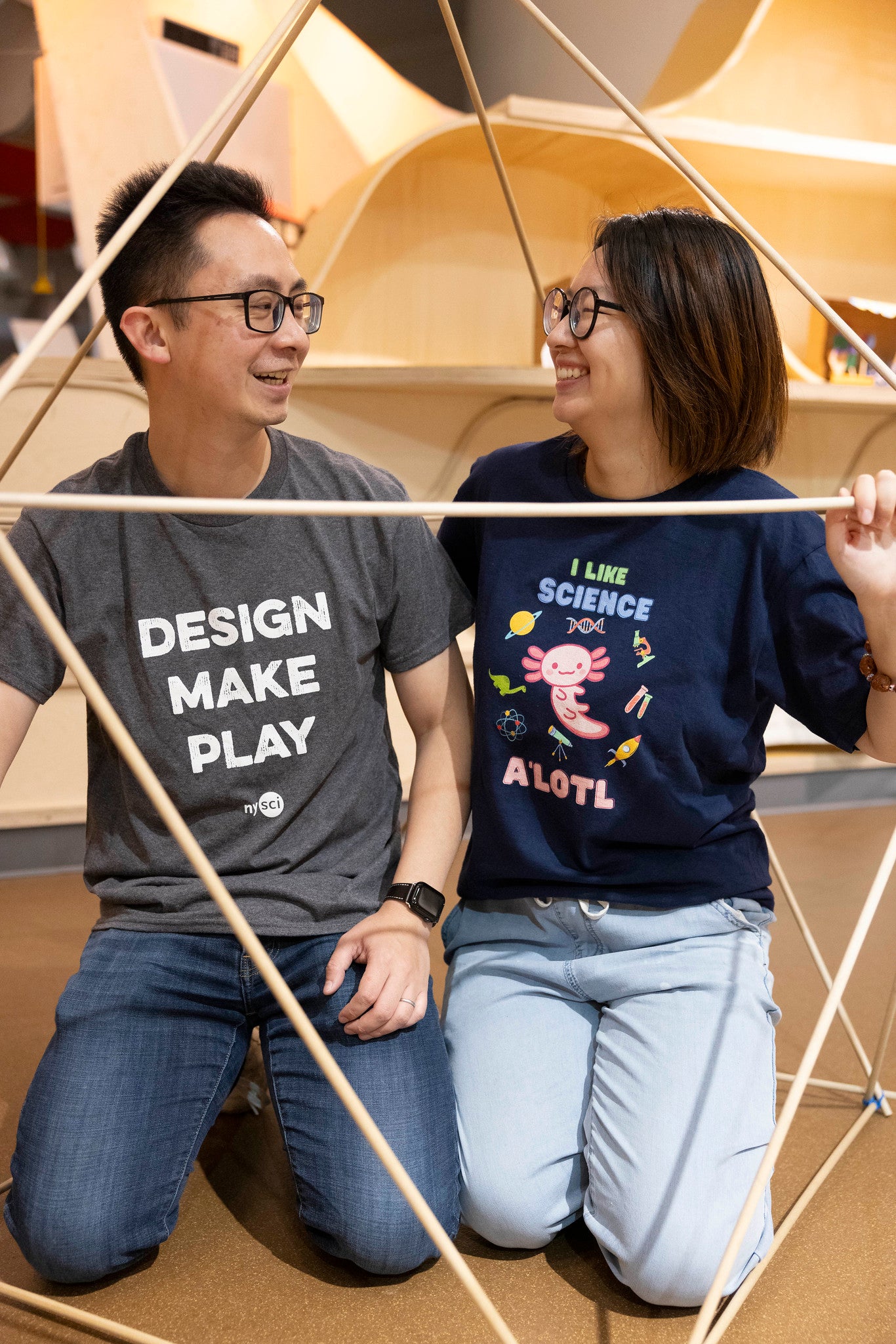 A couple looking at each other. The male has a t-shirt which is gray and says DESIGN MAKE PLAY on it, while the woman has a t-shirt which says I like science a&