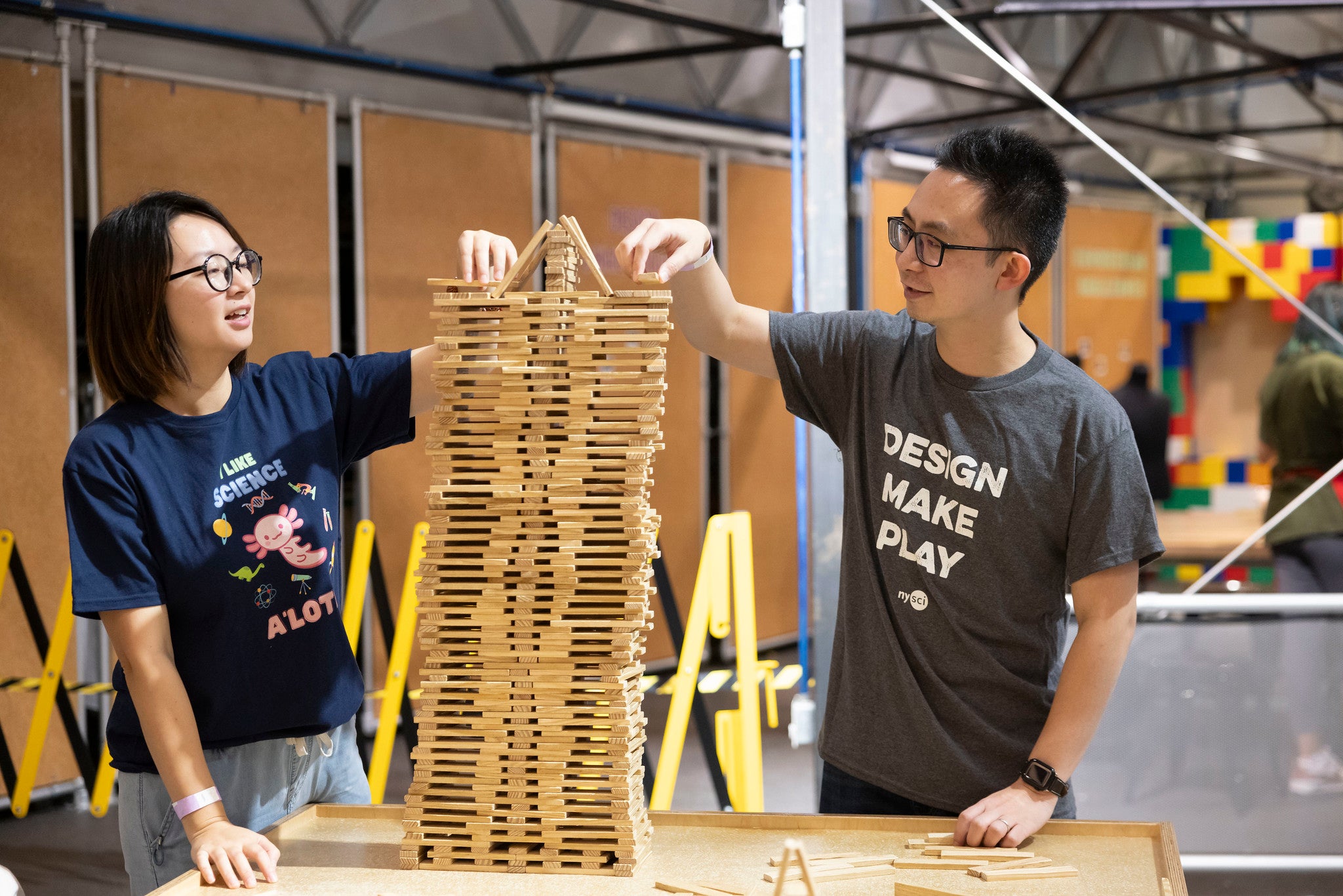 A couple playing together as they stack wooden bricks. The man has a grey t-shirt which says DESIGN MAKE PLAY while the woman has a Navy t-shirt which says I Like Science A&