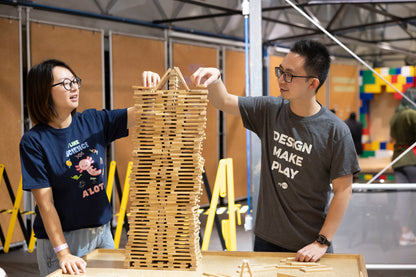 A couple playing together as they stack wooden bricks. The man has a grey t-shirt which says DESIGN MAKE PLAY while the woman has a Navy t-shirt which says I Like Science A&