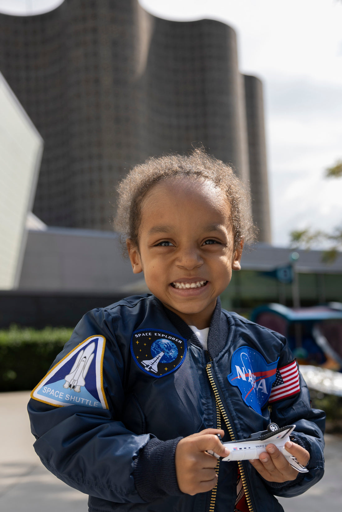 Child looking at camera with a dark blue jacket which has various patches on it, such as a space shuttle, NASA meatball, American flag and another space shuttle going to the earth. There is a gold zipper and the child is outside.