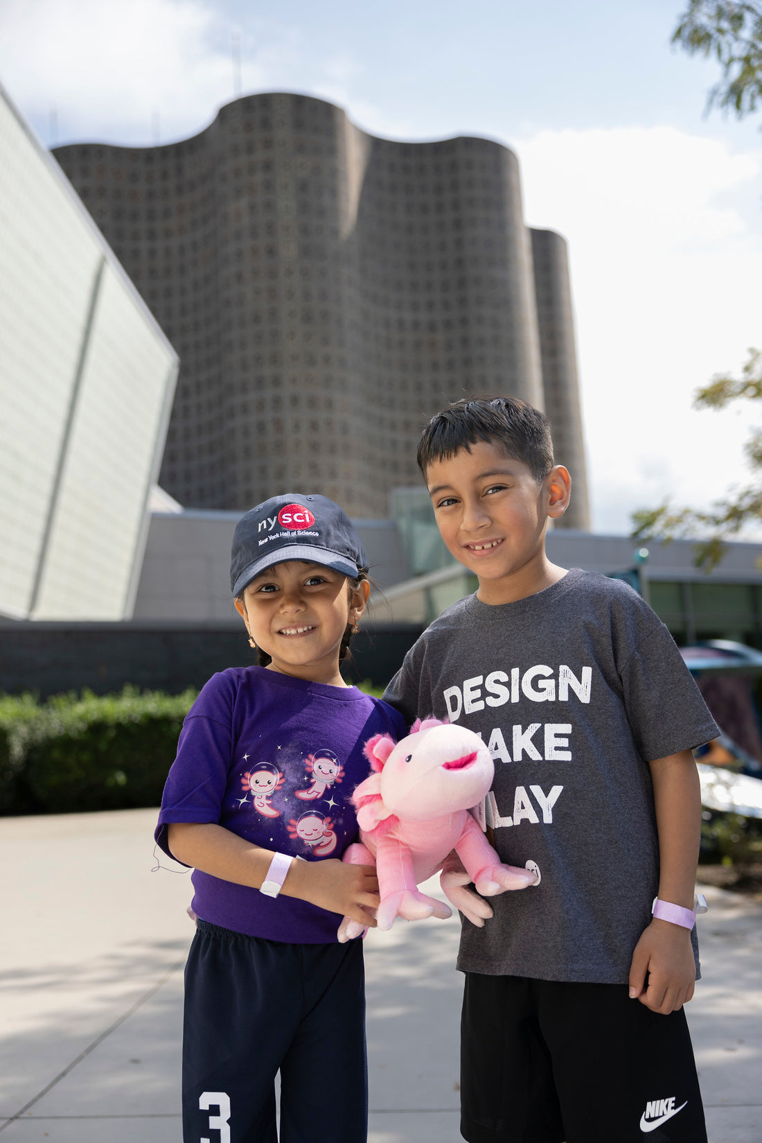 Two children smiling at the camera. One child has a NYSCI baseball cap on which is Navy and has the NYSCI logo. The other child has a gray t-shirt on which says DESIGN MAKE PLAY. THe other child is holding a pink plushie of an axolotl.