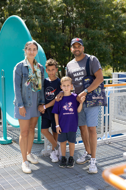 A picture of a father, mother, and two children. They have various shirts on. The father has a grey shirt which says DESIGN MAKE PLAY on it with a blue tote bag while the little boys have a shirt that says astrolotl and the other has the NASA meatball on it. The mother has a varied-colored postcard scarf. They stand and look at the camera, smiling.