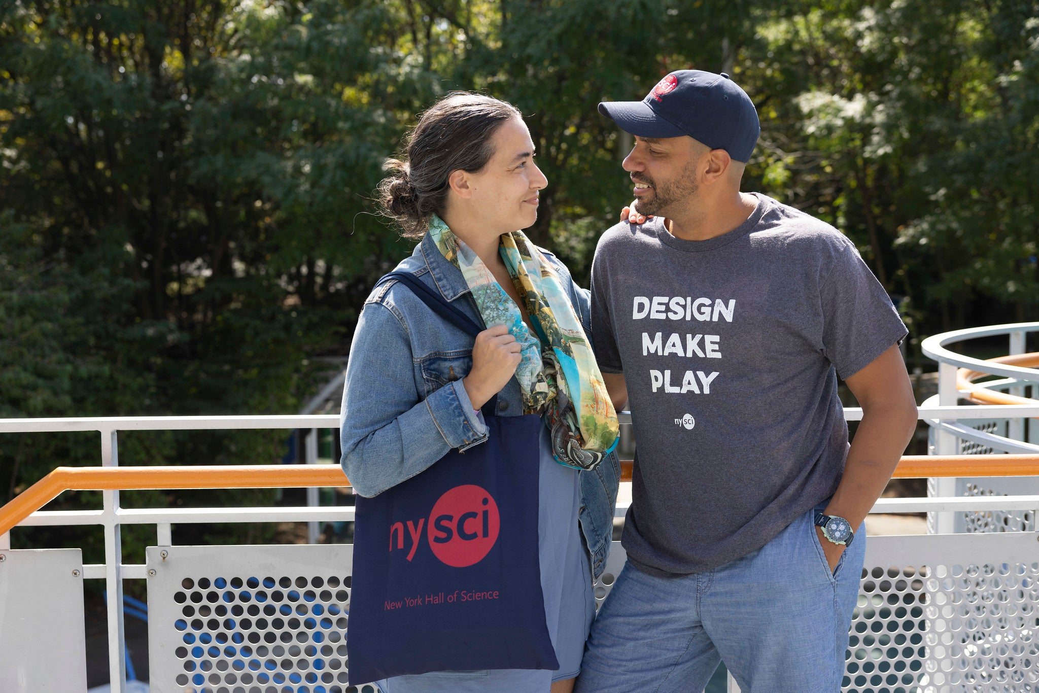 A couple looking at each other. The male has a grey t-shirt on which says DESIGN MAKE PLAY. The woman has a NYSCI logo tote bag.