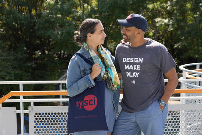 A couple looking at each other. The male has a grey t-shirt on which says DESIGN MAKE PLAY. The woman has a NYSCI logo tote bag.