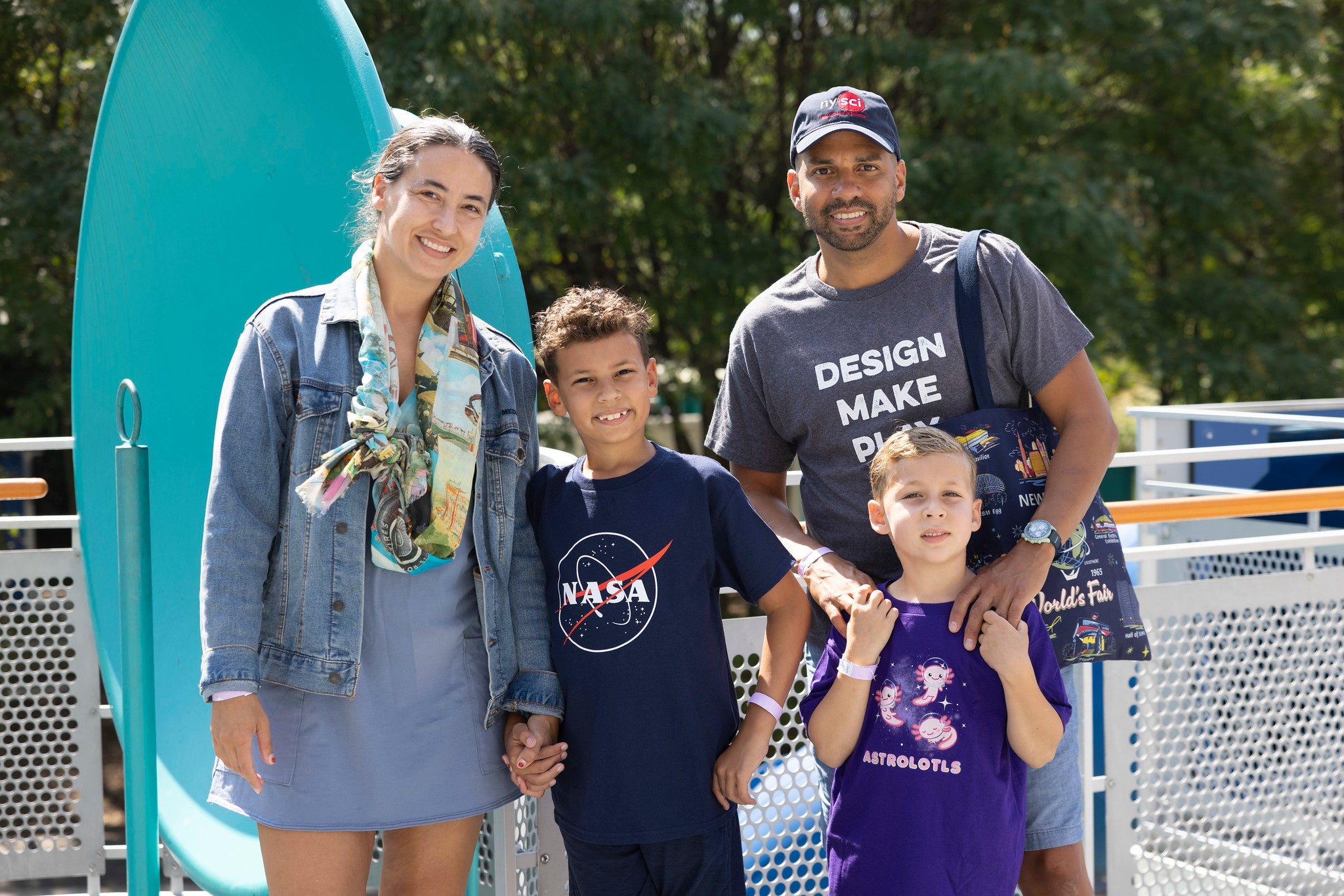 A picture of a father, mother, and two children. They have various shirts on. The father has a grey shirt which says DESIGN MAKE PLAY on it with a blue tote bag while the little boys have a shirt that says astrolotl and the other has the NASA meatball on it. The mother has a varied-colored postcard scarf. They stand and look at the camera, smiling.