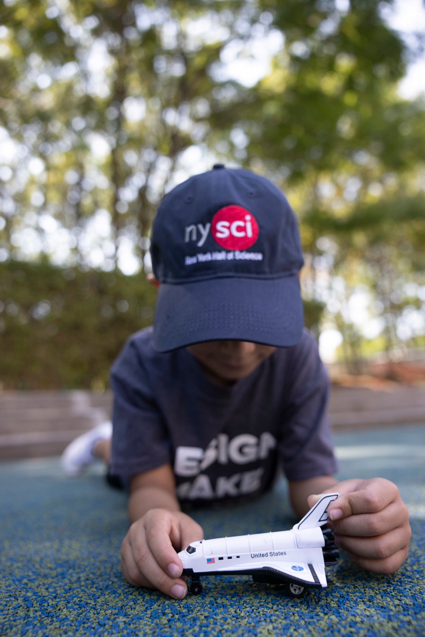 A child laying on his stomach on the ground with a baseball cap which says NYSCI. He is playing with a spaceship toy.