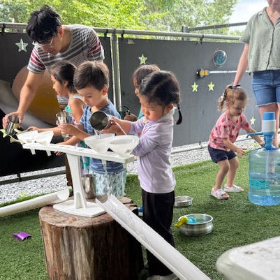 Children playing with water toys outdoors on a grassy area, supervised by adults.