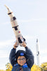 Boy holding up a plush NASA rocket in the park wearing a NASA jacket and hat